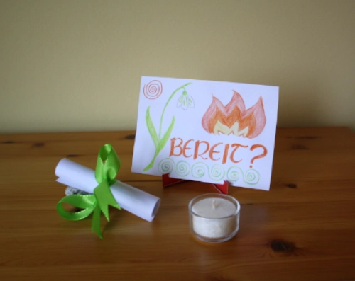 a small DIY stand made from red cardboard with a piece of paper on it. On the paper there are drawing of a snowdrop and a stylised fire, also the question "ready?" in german. In front of the stand there is a tea candle and a small roll of paper held together with a green bow