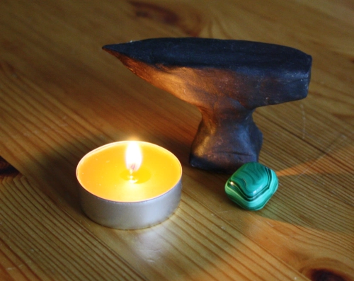 A miniature anvil made from clay and painted brown next to a green semi-precious stone and a burning candle
