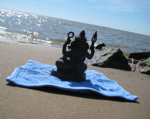 eine kleine Ganeshastatue auf einem blauen Froteelappen am Strand. Im Hintergrund glitzert das Waser im Sonnenschein