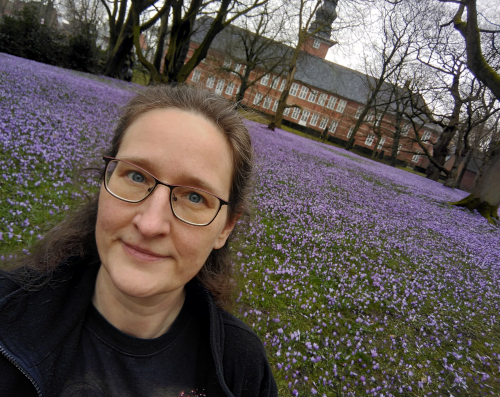 Rückblick März 23: Selfie von Angela vor dem Husumer Schlosspark. Die Wiese hinter mir ist mit unzähligen violetten Krokussen übersät.