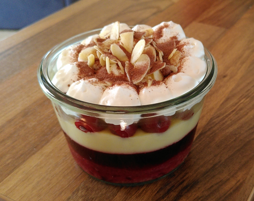 ein Trifle in einem Weckglas. zu sehen sind Pudding-, Gelee-, Kuchen- und Sachneschichte in abwechselnd rot und weiß