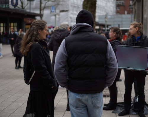 Bestimmung: Ich stehe in der Hamburger Innenstadt und spreche mit einem Mann, der von hinten zu sehen ist. Im Hintergrund stehen zwei Menschen und halten Fernsehbildschirme. Was diese Schirme anzeigen, ist unscharf gestellt, genauso die Gesichter der anderen Personen