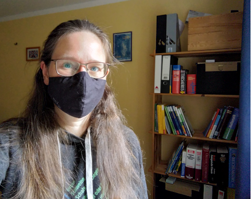 Eine Frau mit FFP2 Maske aus schwarzem Stoff schaut in die Kamera und steht vor einem Regal mit Unterrichtsmaterial und Büchern