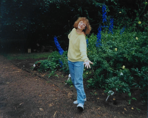 Ein Photo aus den frühen neunziger Jahren. Eine junge Frau mit Dauerwelle, hellgrünem Pullover und mittelblauer Jeans steht mit ausgebreiteten Armen und einem Grinsen im Gesicht vor Blumenrabatten.
