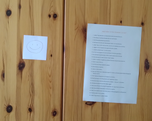 Nahaufnahme von einem Kleiderschrank aus hellem Holz, an der linken Tür klebt ein weißes quadratisches Stück Papier mit einem handgemalten Smiley darauf, an der rechten Tür klebt ein DIN A4-Blatt mit einer Liste