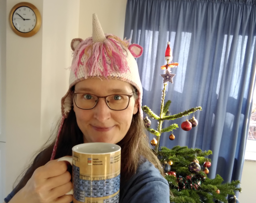 Eine Frau mit einer gehäkelten Einhornmütze, die einen großen Kaffeebecher mit Periodensystemaufdruck in der Hand hält. Im Hintergrund ein Weihnachtsbaum.