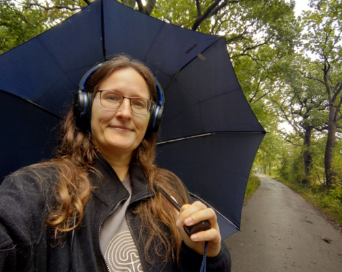 Eine Frau mit Kopfhörern und einem Regenschirm draußen unter Bäumen. Im Hintergrund eine schmale Straße.