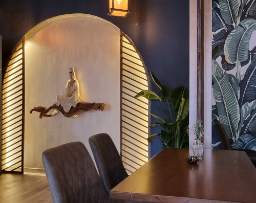 Ein Photo in einem Restaurant, in einer indirekt beleuchteten Wandnische sitzt eine weiße Statue im Schneidersitz. Die Wand ist blau mit einem Blättermuster darauf gemalt.