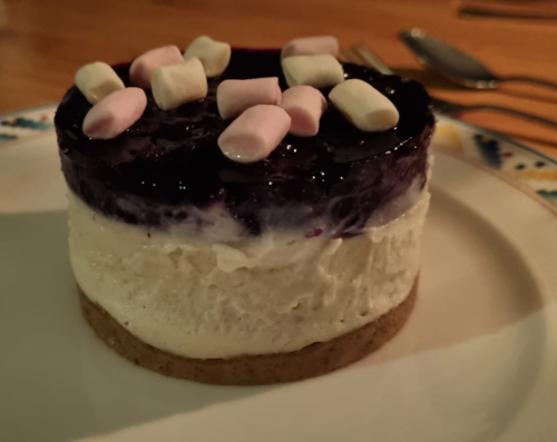 Ein kleiner dreischichtiger Cheesecake auf einem Teller. Die oberste Schichte ist ein Blaubeergelee, darauf liegen noch ein paar Mimimarshmallows