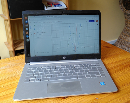 Abiturklausur Analysis: Photo von einem Laptop auf einem Holztisch, im Hintergrund ein Regal mit Lernmaterialien. Auf dem Bildschirm ist die Webseite Geogebra geföffnet