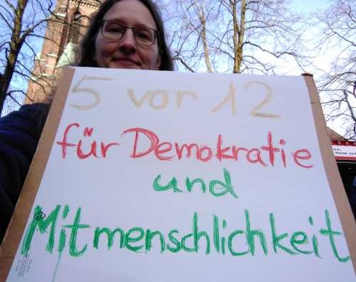 Eine Frau hält auf einem Marktplatz ein Demoschild, darauf steht: fünf vor zwölf für Demokratie und Mitmenschlichkeit