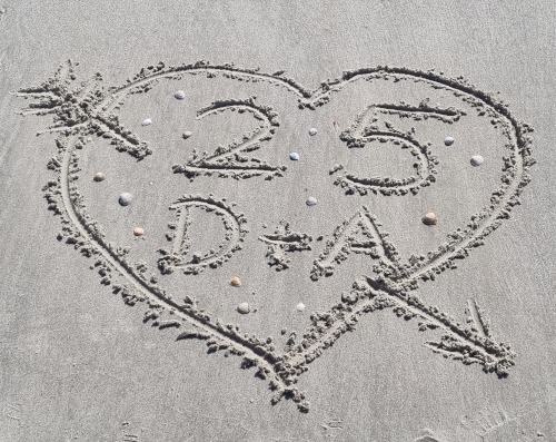 Nahaufnahme von einem Herz, mit dem Finger in feuchten Sandboden geschrieben. Im Herz die Zahl 25 und die Buchstaben D und A, außerdem ein paar Muscheln.