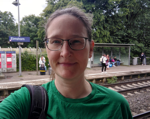 Eine Frau in einem Selfie an einem Bahnhof. Auf dem gegenüberliegenden Gleis ist ein blaues Ortsschild zu sehen, darauf steht: Elmshorn
