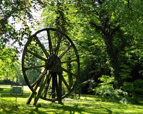 Ein Rad mit ca. 4 m Durchmesser und 12 Speichen in einem Gestell steht in einem sonnendurchfluteten grünen Park. Neben dem Gestellt steht ein Schild auf dem steht: GRuß der Stadt Bottrop Seilscheibe aus der Zeche Prosper