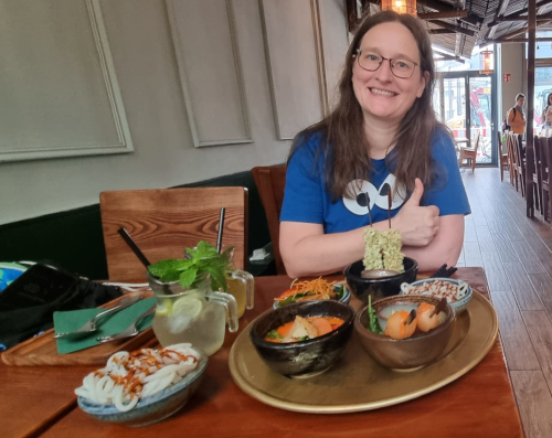 Über 50: Eine Frau mit einem blauen Krümelmonster-T-Shirt sitzt in einem Restaurant am Tisch, grinst in die Kamera und macht einen Daumen nach oben.