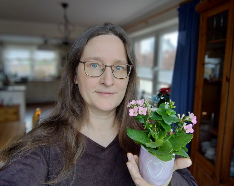 100 Dinge 2024: Nahhaufnahme von einer Frau, der Hintergrund, ein Wohnzimer, ist weichgezeichnet. Inder Hand hält sie eine Topfplanze mit rosa Blüten, der Übertopf ist auch rosa und hat ein kleines Herz aufgeprägt.