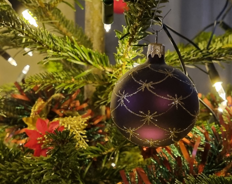 Weihnachten entspannt: Eine lila Kugel mit goldenem Glitzermuster hängt an einem Tannenbaum. Daneben leuchten Lichterketten, außerdem gibt es eine rot-grüne Girlande, die in einer Blumendekoration endet