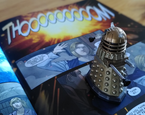 Eine Miniaturfigur eines Daleks steht auf einem aufgeschlagenenen Doktor Who Comic. Am obenren Seitenrand steht das Wort Thooooom vor einer gelben Explosion.