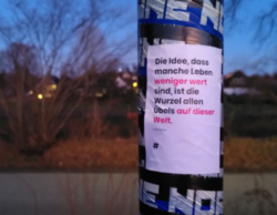 Ein Straßenschildpfahl aus der Nähe photographiert, darauf eine aufkleber mit diesem Text: Die Idee, dass mache Leben weniger wert sind, ist die Wurzel allen Übels auf dieser Welt