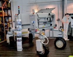 an agricultural robot as a technology example. A light grey machine on four small wheels with many tubes, switches and electronical equipment attached. In the background a shelf containing agricultural museum exhibits, in front of the robot is a stand with leaflets