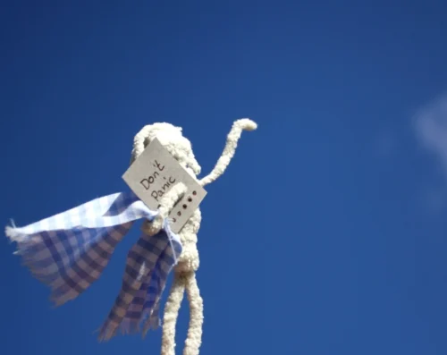 Figur aus Pfeifenputzern, die in der einen Armbeuge ein kleines Handtuch und ein Stück Pappe hält. Auf der Pappe steht: Don't Panic. Den anderen Arm hat sie zum blauen Himmel gereckt.