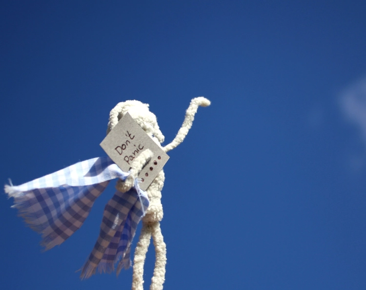 Figur aus Pfeifenputzern, die in der einen Armbeuge ein kleines Handtuch und ein Stück Pappe hält. Auf der Pappe steht: Don't Panic. Den anderen Arm hat sie zum blauen Himmel gereckt.