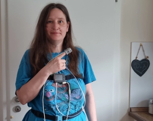 Frau mit einem verschlafenen Gesicht, sie ist mit Gurten und einem elektronischen Gerät verkabelt und hält ihren Zeigefinger hoch, an dem ein weiterer Sensor angebracht ist. sie steht in einer Küche.