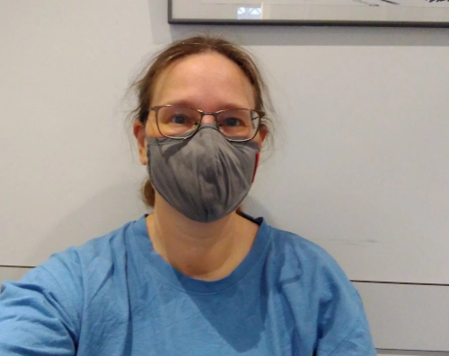 Selfie von einer Frau in einem weiß gestrichenen Flur, sie sitzt und hat eine Maske auf.