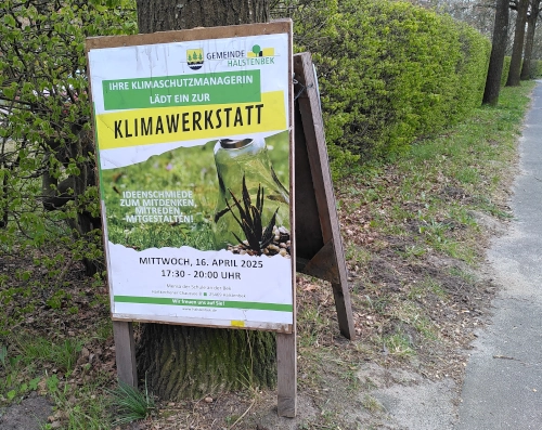 Sandwichaufsteller mit einem Plakat, das zur Klimawerkstatt in Halstenbek einlädt. Das Plakat ist draußen an einem Baum am Straßenrand befestigt.