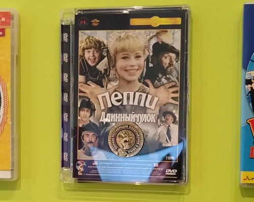 Nahaufnahme einer DVD-Hülle, die an einer Wand hängt. Darauf kyrillische Buchstaben und die Bilder verschiedener Personen, es handelt sich um einen Pipp-Langstrumpf-Film. Pippi sieht blond aus und lächelt.