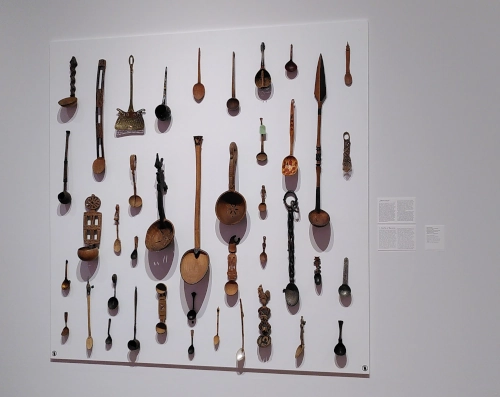 Weiße Wand in einem Museumsraum. An der Wand sind sehr viele sehr unterschiedliche Löffel und Kellen angebracht.