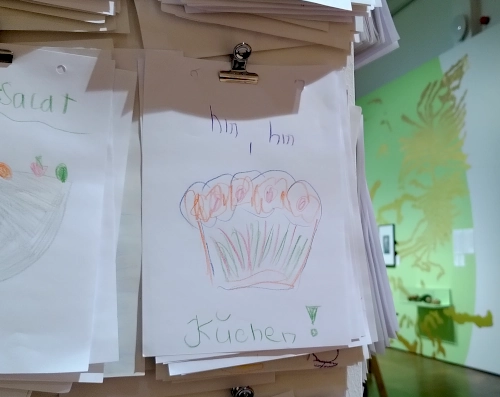 Nahaufnahme von einer Kinderzeichnung, die mit einer Metallklammer an einem Innenraumpfeiler befestigt ist. Darauf ist in buntstift ein Kuchen abgebildet, der Text dazu: Hm, hm, Kuchen!