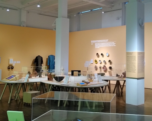 Innenaufnahme eines Ausstellungsraumes in einem museum. An der Wand hängen Teller und Gewänder, auf Tischen stehen verschiedene Gefäße.