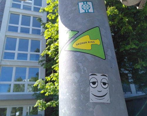 Nahaufnahme von einem Laternenpfahl. Darauf geklebt ein Sticker mit einem lächelnden Cartoongesicht, außerdem ein gelb-grüner Pfeil, der anzeigt, dass hier der Wanderweg "Grüner Ring" verläuft.