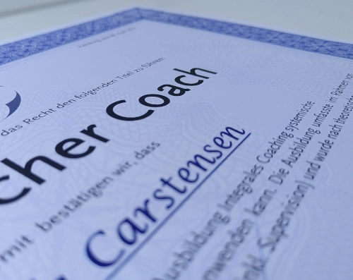 Nahaufnahme einer Urkunde. Das Dokument ist nur zum Teil im Bild, außen verläuft ein violetter Rahmen, die Worte Coach und Carstensen sind zu erkennen.