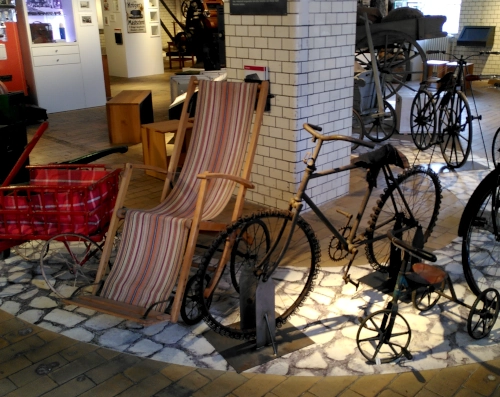 Ein Raum in einem Museum, darin mehrere Fahrzeuge wie Fahrräder, Dreiräder, Handwägen und Rollstühle