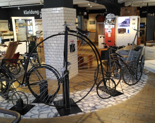 Ein Raum in einem Museum, darin mehrere Fahrzeuge wie Hochräder, Laufräder, und Handwägen