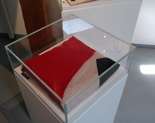 Eine Vitrine, darin eine zusammengefaltete Flagge. Es ist zu erahnen, dass es sich um eine Hakenkreuzflagge handelt, allerdings sind durch die Faltung von dem weißen Kreis nur wenige Prozent zu sehen.