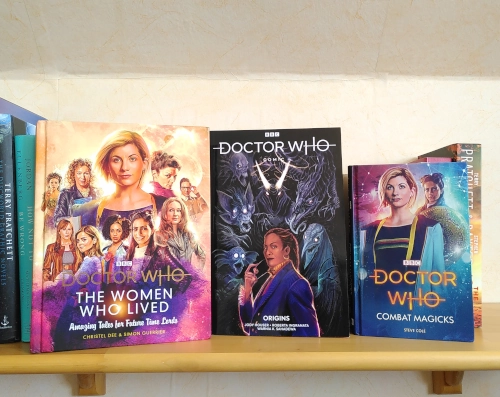 Drei Büchern auf einem Wandbord. Links: docotor Who, the women who lived, in der Mitte ein Comicbuch mit dem Titel Doctor Who, Origins, rechts Doctor Who, combat magicks