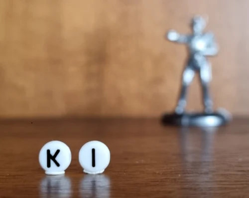 Nahaufnahme von zwei weißen Perlen, die hochkant in einem Regal aus dunklem Holz stehen, auf den Perlen stehen die Großbuchstaben K und I,, sodass auf ihnen der Begriff KI zu lesen ist. Im Hintergrund ist weichgezeichnet eine Roboterfigur zu sehen.