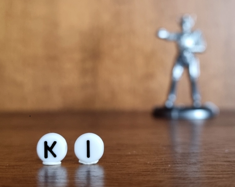 Nahaufnahme von zwei weißen Perlen, die hochkant in einem Regal aus dunklem Holz stehen, auf den Perlen stehen die Großbuchstaben K und I,, sodass auf ihnen der Begriff KI zu lesen ist. Im Hintergrund ist weichgezeichnet eine Roboterfigur zu sehen.