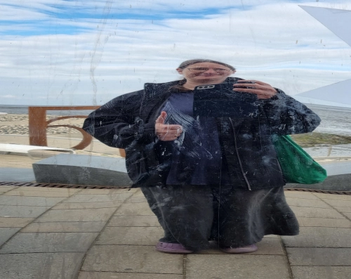 Selfie vom Spiegelbild einer Frau in einem Zerrspiegel. Ihr körper scheint zusammengestaucht, kurz und breit, fast kugelförmig. Sie grinst und hält einen Daumen nach oben. Im Hintergrund bewölkter Himmel und ein Strand.