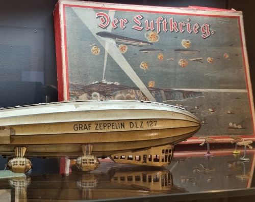 Photo von einem historischen Kriegsspiel namens "Der Luftkrieg" in einem vergilbten Karton. Der Karton steht mit einem Miniaturzeppelin in einer Museumsvitrine.