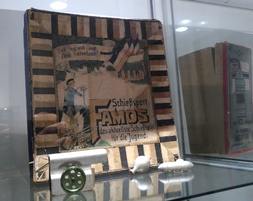Photo von einem historischen Kriegsspiel namens "Schießsport Famos" in einem vergilbten Karton. Der Karton steht mit Miniaturbomben in einer Museumsvitrine.