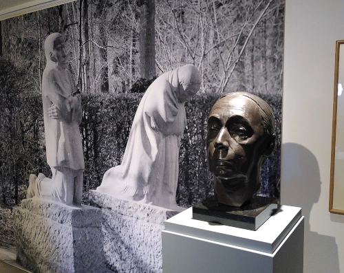 Photo von einer Bronzebüste von Käthe Kollwitz, im Hintergrund an der Wand ein sehr großes Photo, das zwei ihrer Skulpturen zeigt. Die beiden Figuren knien trauernd, der Kopf der Büste schaut ernst.