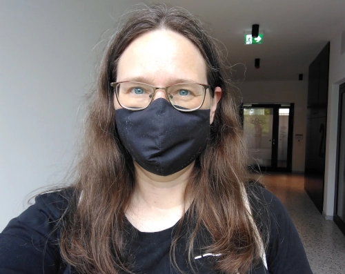 Selfie von einer Frau in einem Flur, sie trägt ein schwarzen langärmeliges Oberteil, eine schwarze Maske und eine Brille und lächelt in die Kamera