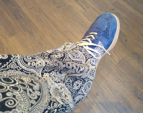 Photo von einem im Sitzen übergeschlagenen Bein, im Hintergrund ein mittelbrauner Fußboden. Das Bein steckt in einer schnörkelig bedruckten Hose in schwarz-gold und der Fuß in einem blauen Schuh