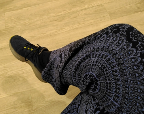 Photo von einem im Sitzen übergeschlagenen Bein, im Hintergrund ein heller Fußboden. Das Bein steckt in einer mandalamäßig bedruckten blauen Hose und der Fuß in einem blauen Turnschuh