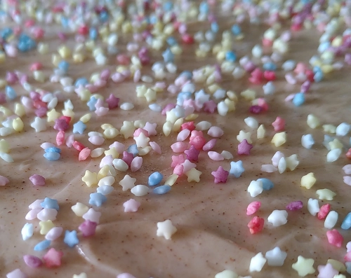 Nahaufnahme von kleinen Sternchen aus Zucker, gelb, rose, blau und weiß auf einen Untergrund aus hellbraunem Zimtzuckerguss gestreut