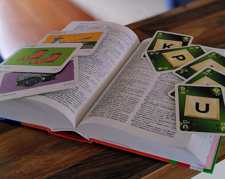Nahaufnahme von einem Wörterbuch Deutsch-Französisch, darauf liegen Karten mit Buchstaben und Karten mit Bildern und den passenden englischen Vokabeln für Stiefeln, Kamera und Flughafen. Der Hintergrund ist weichgezeichnet.